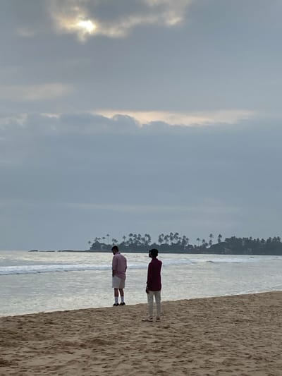 The scene depicts a sandy beach in Dikwella, Sri Lanka, under an overcast sky with a partially obscured bright light source in the upper center. The ocean, featuring gentle waves, occupies the mid-ground. A landmass densely populated with palm trees is visible in the distance, center-right, with a smaller rocky outcrop to its left.
Two adult males stand on the beach, both facing the ocean with their backs to the viewer. The individual on the left, positioned closer to the water, wears a light pink collared long-sleeved shirt, white shorts, dark ankle socks, and dark athletic shoes. The individual on the right stands slightly further inland, dressed in a dark maroon long-sleeved shirt, light-colored long trousers, and dark slide sandals with a white stripe. No direct interaction between the two subjects is observed.