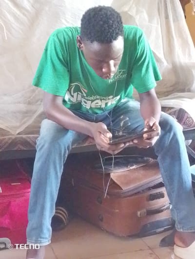 A young Black male, wearing a green t-shirt with "Nigeria Celebrates Sixty-Four" printed on it, and blue jeans, sits on a bed with a mosquito net behind him. He is looking down at a tablet device he holds in his hands, with earphones plugged into it. Two suitcases are stacked on the floor beside the bed. The image includes a TECNO logo in the bottom left corner. The location is identified as Gumel, Nigeria.