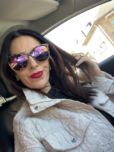 A woman is seated in a car, leaning back against the headrest. She is wearing large sunglasses with purple reflections, red lipstick, and earrings. She has long brown hair and is wearing a beige quilted jacket. Her left hand is holding her hair. The reflection in her sunglasses suggests a cityscape. Through the car window, a building is visible. The location is in Roodt-sur-Syre, Luxembourg.