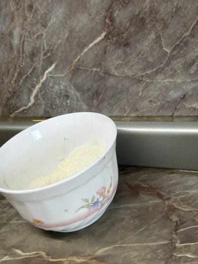 An indoor, close-up, eye-level shot captures a white bowl containing a light-yellow, frothy substance. The bowl, decorated with a floral design, rests on a brown marble countertop. Behind the bowl, a silver metallic backsplash separates the countertop from a darker, patterned marble wall. The scene is devoid of any individuals or discernible activity, suggesting a static, everyday setting within a kitchen or dining area. The location is identified as Crokhovo, Russia.