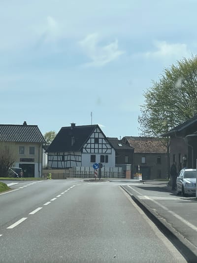 The image shows a street scene in Zülpich, Germany. A half-timbered house with a black roof and white walls with black wooden frames is visible on the side of the road. A blue and white road sign with an arrow pointing to the right is located nearby. Other houses with tiled roofs and modern features line the street. A grey car is parked on the street behind the half-timbered house. The road has white lane markings. A tree is visible on the left side of the image, and the sky is blue with some clouds.