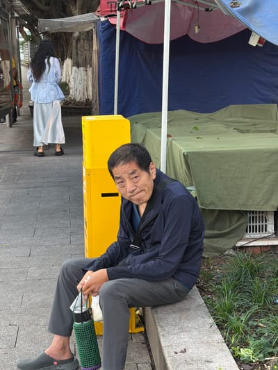 A man sits on a concrete ledge in Longta, China, holding a green bottle cover. He wears a navy jacket and gray pants, with gray clogs on his feet. To his left is a bright yellow utility box. Behind him, a woman in a light blue top and long white skirt walks away, her dark hair flowing. In the background, a market stall is set up with green tarps covering tables, and a blue and pink canopy overhead. The scene appears to be a street in a city.