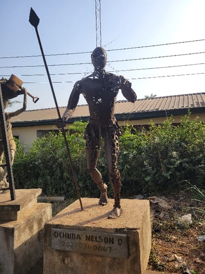The image displays an outdoor scene in the city of San Martín, Colombia, under bright daylight. A prominent dark metal sculpture of a male human figure stands centrally on a rectangular concrete pedestal. The sculpture, appearing to be constructed from welded metal pieces, is depicted in a dynamic, striding pose, with its left hand holding a long, dark spear pointing upwards and to the left. The figure wears a simple wrap around its lower torso. An embedded plaque on the front of the pedestal reads "OCHUBA NELSON O" on the upper line and "202003/0047" below it. To the left, a portion of a second, similar metal sculpture on another concrete pedestal is visible, depicting a figure holding a tool resembling a shovel. The background features a low-rise building with a brown corrugated metal roof, partially obscured by green bushes and dense foliage. Overhead, a clear sky reveals multiple power lines and the upper section of a tall metal lattice tower on the right. The ground around the pedestals is unpaved dirt with scattered debris and sparse vegetation.