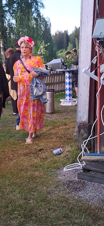 The image depicts an outdoor scene in Salpakangas, Finland. A woman in the foreground is wearing a vibrant pink and orange floral dress and a floral crown. She is holding a clear plastic cup in her left hand and a denim jacket over her right arm. She is also wearing light-colored sneakers. To her right, on the grass, is a discarded aluminum can. In the background, there are several other individuals, a table with a blue and white checkered pattern, a wooden fence, and trees. The setting appears to be a gathering or an event.