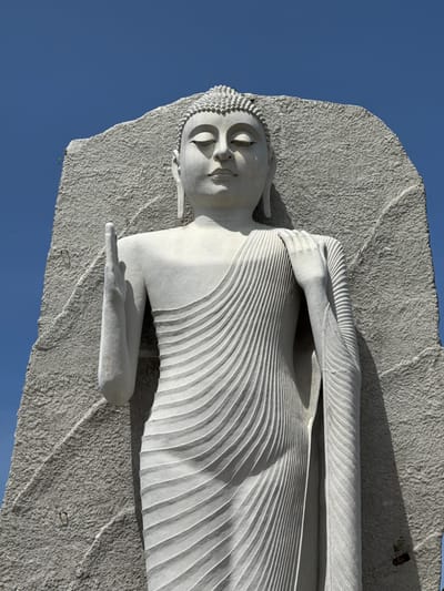 The image shows a close-up, low-angle shot of a large, white Buddha statue carved from stone, set against a clear blue sky. The statue is depicted standing, with its eyes closed and a serene expression on its face. Its right hand is raised with the palm facing outwards, in a gesture of blessing or protection, while its left hand rests on its shoulder, draped with intricate, flowing robes. The stone background behind the statue is rough and textured, contrasting with the smooth finish of the sculpture. The statue is located in Dondra, Sri Lanka.
