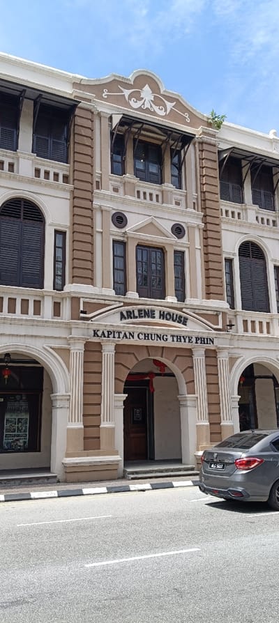 The image showcases a three-story building, the "Arlene House - Kapitan Chung Thye Phin". Its facade is adorned with light-colored pillars and moldings, contrasting with brown window frames and accents. Shutters cover most windows. A car is parked in front of the building, partially obscuring the entrance. The scene is captured under bright daylight, with clear skies above.