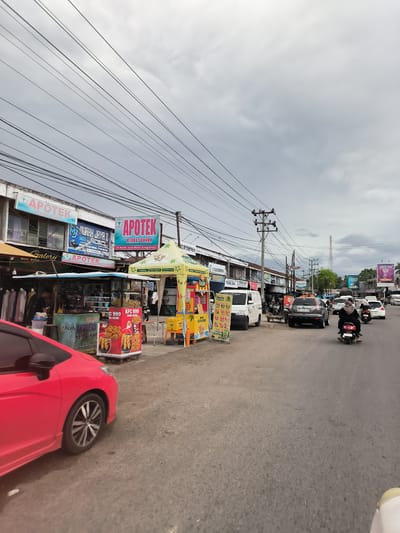 The image presents a busy street scene in Grong-Grong, Indonesia, under an overcast sky. In the foreground on the left, a red car is partially visible, either parked or in motion. Adjacent to the road, a row of commercial buildings lines the street, featuring various shops and businesses.

Prominently displayed are multiple signs for "APOTEK BERKAT BUNDA," with one sign specifying its location as "Jl. Banda Aceh Medan Grong-Grong." Other visible business names include "AURAH JAYA 2," "Galery," "KALIFORNIA," "TRINA JAYA," and an "Aceh AKADEMI" sign indicating student enrollment.

On the sidewalk, several street vendors operate under vibrant yellow and green tents. One stall features "KFC 999" branding, displaying fried chicken and other items. Another stall offers drinks and snacks, including "matcha," with text like "Fresh Brew" and "Kopi Aceh." A person can be seen working at one of the stalls.

The paved road in the mid-ground shows moderate traffic, including a white van, a grey SUV with license plate "BL 1199 PR," a white sedan, and several motorcycles and scooters. One rider on a scooter is visible wearing a headscarf.

The sky is a uniform grey, indicating cloudy weather. A dense network of black utility lines and cables crisscrosses overhead, supported by tall utility poles. In the distance, a radio or telecommunications tower stands against the horizon, and billboards, including one for "VEEVA," are visible further down the street. The scene depicts a lively commercial area with ongoing daily activities.