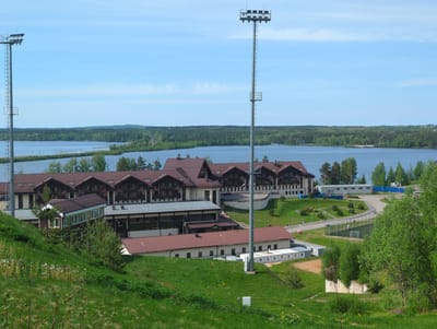 The image captures a wide-angle view of a lakeside landscape, featuring buildings and recreational facilities in what can be identified as Toksovo, Russia. In the foreground, a grassy hill slopes down to reveal several structures. The most prominent is a multi-story building complex with a brown roof and dark brown balconies, potentially a hotel or resort. A long, one-story building with a red roof is also visible in the foreground. A tall metal lighting tower stands between the camera and the complex. In the background is a body of water, likely a lake, with forested land visible on the opposite shore. A small soccer field is visible in the lower right corner. The sky is clear and blue.