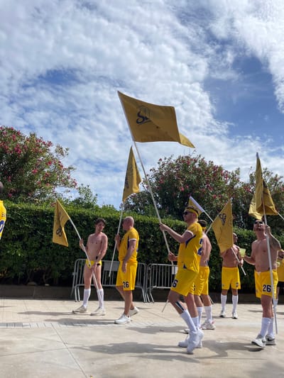 The image depicts a group of men holding yellow flags with a dark blue logo on them, standing in front of a lush green hedge with pink flowering bushes. The men are wearing yellow shorts and some are shirtless, others are wearing yellow jerseys. Several of the shorts have the number "26" visible. The flags appear to be carried on white poles. In the background, there is a metal fence and a cloudy blue sky. The scene suggests an outdoor event or parade. This image was taken in Sitges, Spain, likely during a summer festival or sporting event.
