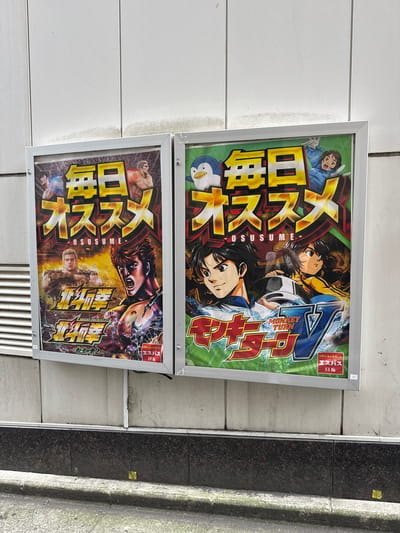 Two framed posters are mounted on a textured white wall outdoors. The poster on the left is primarily dark red, yellow, and brown, featuring animated characters with muscular physiques and intense expressions. The text "毎日 オススメ" (Mainichi Osusume), meaning "Recommended Daily", is prominently displayed in large yellow Japanese characters across the top of the poster. Below this, smaller text reads "OSUSUME". The bottom half of the poster displays Japanese characters that appear to be the title of a game or series, with the words "SMART PACHISLOT" and "北斗の拳2" (Hokuto no Ken 2) visible. The poster on the right has a bright green background with yellow and red lettering. The text "毎日 オススメ" is also displayed in large yellow Japanese characters at the top, with "OSUSUME" below it. This poster features animated characters with larger eyes, including a penguin figure and several human characters. The bottom of the poster contains Japanese text, "MONKEY TURN V", and the words "パチンコ&スロット" (Pachinko & Slot). Both posters are framed in silver metal. A dark gray, textured base runs along the bottom of the wall. The ground in front of the wall is paved and appears to be a street or sidewalk with some moss and debris. The overall setting suggests an outdoor advertising space in a Japanese city. Based on the context of "Pachinko & Slot" posters and the general aesthetic, this scene is likely located in Shinjuku, Japan, a district known for its entertainment and gaming establishments.