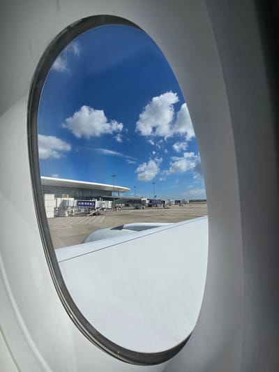 This image, captured from an airplane window in Bao'an District, China, shows a view of an airport tarmac on a clear day. The sky is a vibrant blue with scattered white clouds. In the foreground, the white surface of the airplane wing is visible. Beyond the wing, the airport terminal building is visible with jet bridges extended towards parked aircraft. Several jet bridges are connected to the terminal, and some appear to be empty. A banner with Chinese characters is visible on one of the jet bridges. The tarmac is paved, and there are markings on the ground indicating parking spots for aircraft. Lights and poles are also visible on the tarmac. The scene suggests an active airport environment under a bright sky.