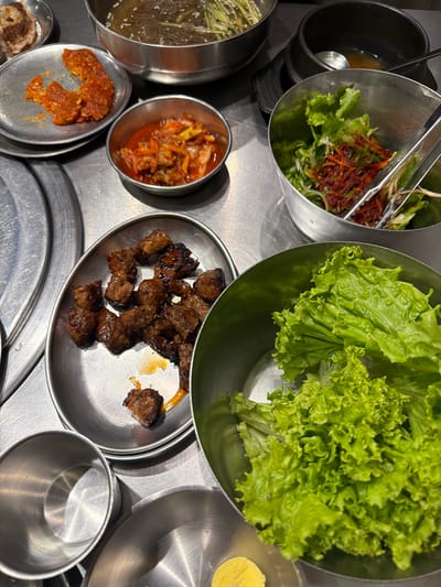 A close-up overhead shot shows a table laden with Korean barbecue dishes in what appears to be a restaurant in Cebu City, Philippines. In the foreground, a large stainless steel bowl is filled with fresh, vibrant green lettuce leaves, their ruffled edges catching the light. To the left, a smaller metal dish contains bite-sized pieces of grilled meat, some glistening with rendered fat and a hint of reddish sauce. Further back, a metal bowl holds a serving of kimchi, its bright orange color contrasting with the other dishes. Another metal bowl contains a salad of shredded vegetables and lettuce, tossed with a reddish sauce and held by silver tongs. A large silver bowl in the upper left contains a noodle dish, possibly naengmyeon. The table surface is a brushed metal, typical of Korean barbecue restaurants. The overall impression is one of a meal ready to be enjoyed, with a variety of fresh ingredients and cooked meats.