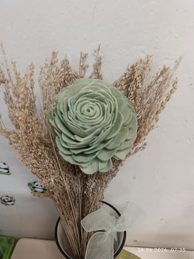 A close-up shot captures a pale green, rosette-shaped dried flower placed in the center of a bouquet of dried wheat stalks. The wheat stalks are a light brown color and form a backdrop for the flower. A light green, sheer ribbon is tied in a bow around the vase, which is partially visible at the bottom of the image. The background is a textured white wall, with some subtle specks and imperfections visible. To the left of the bouquet, on the wall, there are a few small, cartoonish panda stickers. The lighting suggests it is daytime, with a soft, diffused light. A timestamp at the bottom right of the image reads "26.06.2026 07:35". The overall impression is one of natural beauty and simple decoration, with a touch of whimsy from the panda stickers. The setting appears to be indoors, likely a room or a display area.