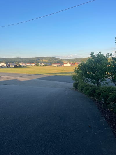 A wide shot shows a rural landscape with a clear blue sky. In the foreground, a dark asphalt path curves to the right. Beyond the path is a field of dry grass, and further back, a cluster of residential buildings with red and white roofs. Behind the buildings, a tree-covered hill rises, and in the distance, more rolling hills are visible. The scene appears to be in the city of Cham, Germany.