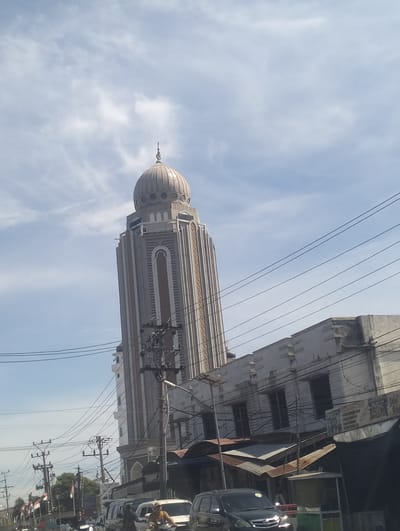 The image depicts a view of a tall, modern mosque minaret topped with a dome, situated in a city in Indonesia, likely Barat Yaman. The minaret, with its distinctive patterned facade and arched windows, dominates the upper portion of the frame. In the foreground and midground, a street scene unfolds with various elements characteristic of an urban environment. Several utility poles with numerous wires are prominent, indicating the presence of electrical infrastructure. A low-rise building with a textured, light-colored facade is visible adjacent to the mosque, its roofline partially obscured by the minaret. Several vehicles, including cars and motorcycles, are present on the street, suggesting traffic. People are seen on motorcycles and near a stall on the right side of the image. Flags, possibly national flags, are displayed on poles on the left side of the street. The sky is partly cloudy, with a clear blue expanse interspersed with white clouds, indicating daylight. The overall impression is of a bustling city street with a significant religious structure.