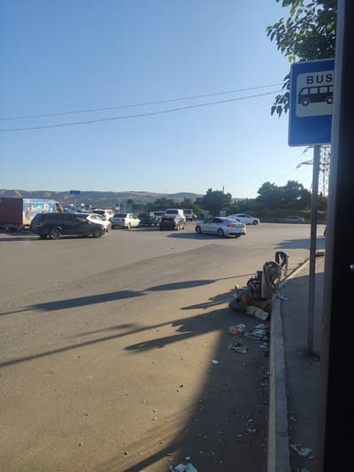 The image captures a street scene in the city of Motodrom YS, Azerbaijan, with a focus on a road and its surroundings. A blue bus stop sign is prominently displayed on the right side of the frame, indicating the location of a bus stop. The road is paved and appears to be a busy thoroughfare, as evidenced by the presence of several cars and a truck. The cars are of various makes and models, suggesting a typical urban traffic flow. The truck, positioned on the left, might be a commercial vehicle, possibly involved in transportation. The environment is bathed in the warm glow of what appears to be late afternoon sunlight, casting long shadows across the road. The sky is clear and blue, indicating fair weather. In the background, distant hills and trees are visible, suggesting a landscape that combines urban elements with natural scenery. The presence of litter along the curb on the right side of the road points to urban activity and the need for waste management. There are no people clearly visible in the image, making it a snapshot of the urban environment in the absence of direct human interaction. The overall impression is one of a functional, albeit somewhat untidy, city street.