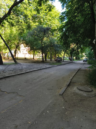 This is a vertical, eye-level, medium shot of a paved road passing through a residential area lined with trees and some buildings in Sokolivske, Ukraine. The road is dark gray and appears to be somewhat worn, with cracks and a faded yellow line visible in the foreground. On the left side of the road, there is a dirt and sand area with several trees and a glimpse of a building with a fence behind them. On the right side, there is a sidewalk with a manhole cover and more vegetation. Two cars are visible in the distance, one parked and one driving away. The scene is dominated by the lush green foliage of the trees, which casts shadows on the road, suggesting it is daytime. The lighting indicates it might be late afternoon or early morning, with sunlight filtering through the leaves. There are no people visible in the frame. The overall impression is of a quiet, somewhat neglected urban or suburban setting during a pleasant season.