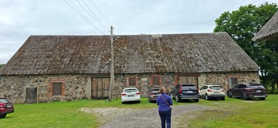 The image captures a wide exterior view of a weathered, stone-built barn with a corrugated, moss-covered roof under an overcast sky. The barn features large wooden double doors in the center, a smaller wooden door on the left, and boarded-up window openings with red brick surrounds. Several cars are parked on the gravel and grass in front of the barn, including a red SUV on the far left, a white hatchback, a dark SUV, a dark sedan, and a light-colored SUV. A woman in a blue jacket and dark pants stands with her back to the camera, facing the barn. The scene is set in Kohatu, Estonia, with a large green tree visible on the right side of the frame. A wooden utility pole with electrical wires runs vertically through the image, near the center. The overall impression is of a rural, somewhat aged agricultural setting.