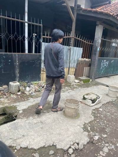 The image shows a young man walking away from the camera on a cracked concrete sidewalk. He is wearing a dark grey long-sleeved shirt and brown pants. The man is wearing black sandals. He is walking past a house with a black metal gate and a broken concrete drain. There is a metal bucket on the right side of the image. The location is on a street in Baleendah, Indonesia at the coordinates -7.021, 107.673.