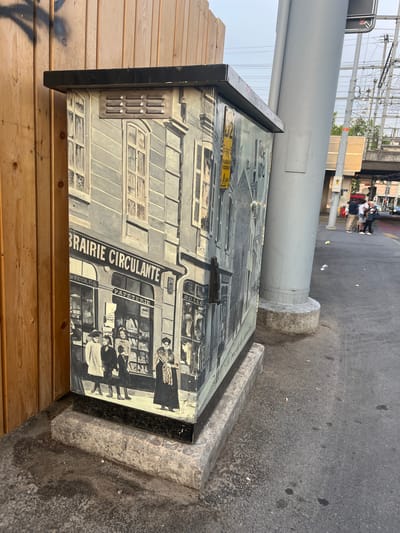The image depicts a mural painted on the side of a utility box. The mural is rendered in black and white and features a detailed scene of a street in Geneva, Switzerland, likely from the early 20th century.

The subjects of the mural are people and buildings. Several groups of people are depicted, some standing on a sidewalk and others inside what appears to be a shop. They are dressed in period clothing, suggesting the scene is historical. The buildings are tall and ornate, with many windows, and a streetcar is also visible, reinforcing the historical setting. A sign on the building reads "PRAIRIE CIRCULANTE" and "PAPETERIE", indicating a shop selling stationery. Another sign appears to say "ÉCOLES".

The scene captures a bustling urban environment. The lighting suggests it is daytime, though the sky is overcast, as seen in the upper portion of the original image where power lines and a large concrete pillar are visible. The utility box is placed on a sidewalk next to a road. The concrete base of the utility box and the surrounding asphalt ground are visible. There are some small white pieces of debris scattered on the ground.

Notable details include the artistic style of the mural, which is realistic and detailed, and the specific signage which provides clues about the location and time period. The overall impression is one of historical preservation and public art.