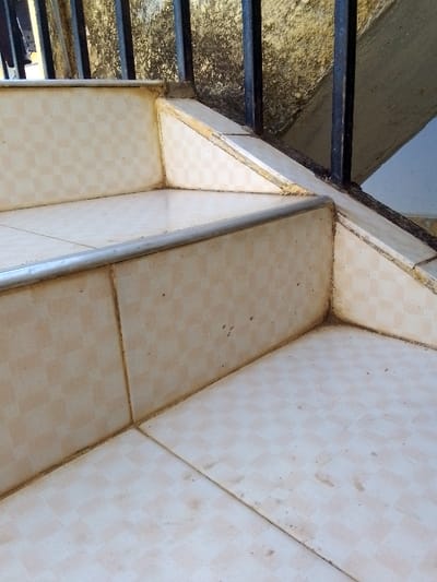 The image depicts a close-up, upward-angled view of a section of an outdoor tiled staircase located in Kalimoni ward, Kenya. The stairs feature light beige or cream-colored square tiles with a subtle checkered pattern on both the treads and risers. A silver-colored metallic strip functions as stair nosing on the leading edge of the visible tread. Black vertical metal balusters, part of a railing system, are anchored into the tiled stairs and extend upwards. The tiled surfaces, visible grout lines, and the rough-textured, discolored wall behind the railing exhibit accumulated dirt, dust, and dark stains. No individuals, specific actions, or interactions are present in the frame.