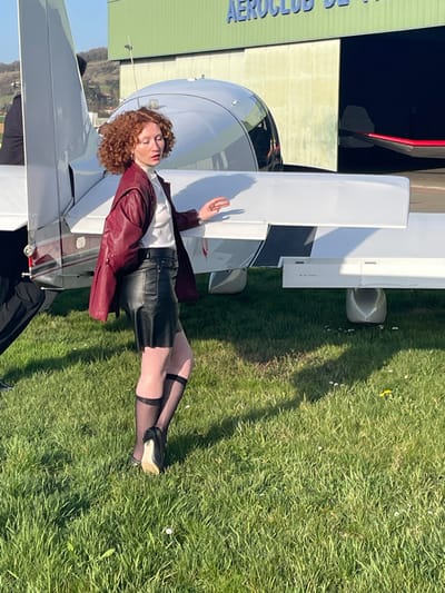 The image displays a female subject posing on a grassy airfield, identified by a hangar sign reading "AEROCLUB DE LYON." This airfield is located in the vicinity of Reventin-Vaugris, France.
The central figure is a female with reddish-brown curly hair and a light complexion. She is positioned facing left with her head slightly turned towards the camera, her eyes partially closed. She wears a white turtleneck shirt, a dark red leather jacket, a black leather mini-skirt, sheer black stockings featuring two horizontal bands above the knee, and black high-heeled shoes. Her right hand is slightly raised with fingers extended.
Behind the female subject, a white single-engine light aircraft is parked on the grass. Its tail section, right wing, and a portion of its cockpit are visible. In the background, a large green hangar with a partially open dark bay displays the "AEROCLUB DE LYON" signage. A segment of a red aircraft is visible inside the hangar's open bay. A second individual, appearing male, is partially visible on the far left, positioned behind the tail of the white aircraft, wearing dark trousers and a dark jacket.
The ground consists of green grass interspersed with small white and yellow wildflowers. Hills are present in the distant left background under a clear blue sky. Shadows indicate a low sun angle.