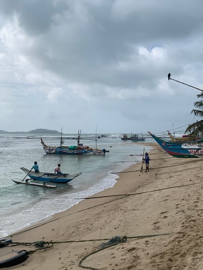 In Mirissa, Sri Lanka, a cloudy day unfolds at the beach with several fishing boats pulled ashore. In the foreground, a sandy beach is lined with ropes and two tires. Closer to the water, a man stands on the shore holding a pole, while another man, seated, prepares to row a small, narrow boat that is partially in the water. In the background, more fishing boats are visible in the shallow, calm water, with one larger, colorful boat adorned with writing. A distant island is visible on the horizon. Above, a bird perches on a pole. The overall scene depicts a tranquil, overcast day by the sea in a coastal town.