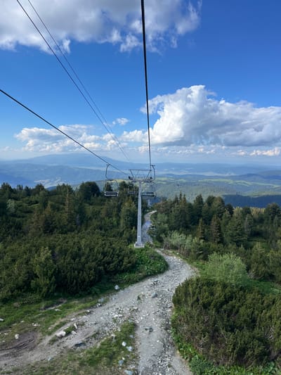 This image captures a scenic view from a ski lift in Panichishte, Bulgaria. The scene is dominated by a vast expanse of lush green pine forests that cascade down a mountain slope. A gravel path winds its way alongside the trees, indicating a trail or service road.
In the foreground, the ski lift is a prominent object. Two empty chairs hang from a cable, suspended in the air, ready to transport unseen individuals. The cable extends upwards and to the left, disappearing into the bright blue sky dotted with fluffy white clouds.
The background reveals a breathtaking panorama of layered mountain ranges fading into a hazy distance. The valley below appears green and somewhat cultivated, suggesting a rural or natural landscape.
The time of day appears to be daytime, with clear blue skies and sunlight illuminating the scene. The weather seems pleasant, with scattered clouds suggesting fair conditions. The overall impression is one of natural beauty and the potential for outdoor recreation. No visible text is present in the image.
