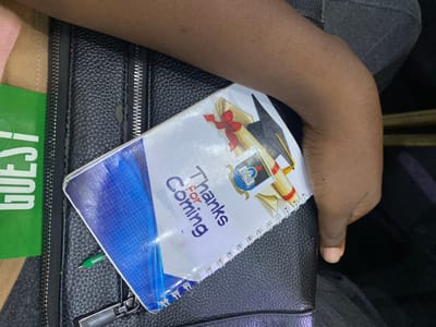 In Ikoyi, Nigeria, a close-up shot shows a person's hand resting on a black, textured bag. On the bag, a white spiral notebook is placed, its cover featuring a graduation cap, a diploma scroll with a red ribbon, and text that reads "Thanks For Coming." A small green pen is tucked into the notebook. To the left of the notebook, a green card with the word "GUEST" printed in white is visible. The background is dark, suggesting an indoor setting.