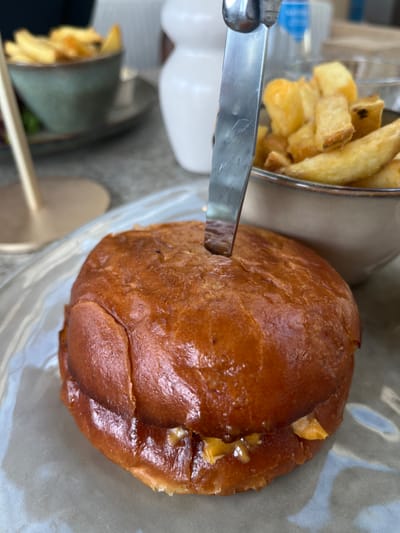 The scene, set in Overveen, Netherlands, features a close-up of a single burger on a light gray plate. A reflective metal knife is vertically inserted into the center of the burger's glossy, golden-brown bun. Orange-yellow melted cheese is visible extruding from the lower portion of the burger. Adjacent to the burger, a beige ceramic bowl contains golden, thick-cut french fries. In the blurred background, additional restaurant elements are present, including a second bowl of food, a white ceramic dispenser, and a clear drinking glass. No individuals are depicted.