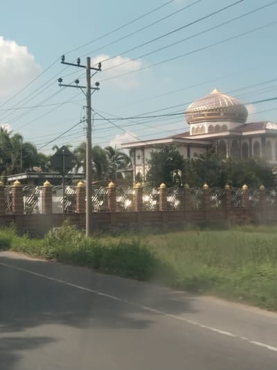 The image depicts a roadside view of a building with a large dome, possibly a mosque, located in Payameuneng, Indonesia. The building features white walls with decorative arches and a prominent golden dome. It is surrounded by a decorative fence with brick pillars topped with golden finials and intricate metalwork. Tall palm trees and other lush vegetation are visible behind the fence, framing the building. A utility pole with multiple wires stands in the foreground, crossing the frame. The scene is captured from the perspective of a moving vehicle, as indicated by the blur of the foreground and the presence of a road with a white lane marking. The sky is blue with scattered clouds. The image appears to be taken during daylight hours.