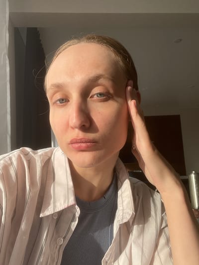 A fair-skinned person with light hair and green eyes is positioned in the foreground, with their left hand touching their forehead. The individual is wearing a gray ribbed tank top covered by a striped white and light pink button-down shirt. Light from an unidentified source casts shadows on their face and the surrounding environment. The background includes a section of a light-colored wall and a dark wooden or brown surface that could be a door or piece of furniture. There is also a metallic object, possibly a handle or rail, visible at the top of the frame. No indicators can confirm the location as Sepych, Russia.
