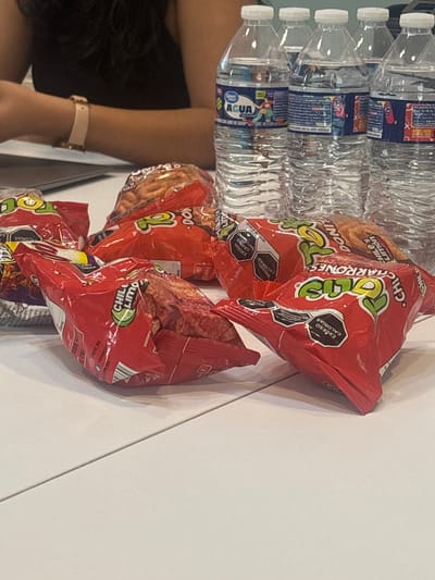 The image shows a partial view of a person's left arm wearing a watch, alongside a stack of water bottles and several bags of Donitas Chile y Limón-flavored wheat snacks. The water bottles appear to be "Great Value" brand. The snack bags have nutritional information printed on them, including calorie and fat content. The text on the bags suggests the product is made in Mexico. The image also indicates the snacks are available for purchase at a "COSTCO" location, based on the text near the bottom of one of the bags. No other specific details about the location in Buenavista, Mexico, are visible in the image.