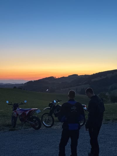 The image depicts a twilight scene with a clear gradient sky transitioning from deep blue at the top to orange and yellow hues near the horizon. In the foreground, two individuals, silhouetted against the dusk, stand near two dirt bikes. One bike is predominantly red and white, while the other is black. The individuals are wearing dark clothing. Behind them, rolling hills covered in green grass and trees recede into the distance, with layers of mountains visible further back. The ground in the immediate foreground is covered with gravel. The location is identified as St. Peter, Germany.