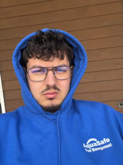 A medium shot photograph of a man with dark curly hair, glasses, and a beard is positioned in a blue hoodie with the word "Aquasafe Pool Management" printed on the right side. He is wearing glasses that reflect the surroundings. The background is a wooden wall. The image appears to be taken outdoors in daylight. The exact location in Edison, United States of America, is not identifiable from the provided image.