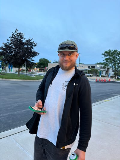 A man stands on a sidewalk in Morton Grove, Illinois, holding a smartphone in his left hand and a can in his right. He has fair skin, a beard, and is wearing a baseball cap with sunglasses on top, a black hooded jacket over a gray t-shirt, and black pants. The man is facing towards the camera at an oblique angle. Behind him is a street with cars and trees. Further in the background, a "Fifth Third" bank branch is visible, along with traffic cones. A dark-leafed tree is located to the left of the man. The sky is overcast.