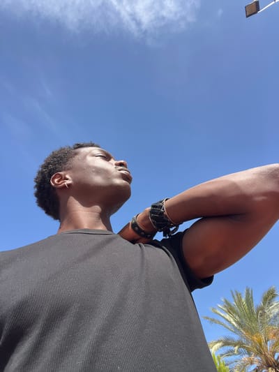 A low-angle shot shows a Black man from the chest up, looking up towards a clear blue sky. He wears a black ribbed t-shirt and has his right arm bent and resting behind his head. He has dark, curly hair and a thin hoop earring in his left ear. He is wearing several bracelets on his right wrist, which appear to be made of leather and metal. Sunlight highlights his skin and the contours of his face and arm. In the upper right corner of the image, a light fixture with a square brown shade is visible, attached to a white pole against the sky. In the lower right, the fronds of a palm tree are visible. The overall scene suggests an outdoor setting, possibly in a warm climate. The presence of the palm tree and the light fixture is consistent with an urban outdoor public space, like a park or plaza, potentially in a Mediterranean city. Given the location of Sant Adrià de Besòs, Spain, it is plausible that this image was captured in one of its public recreational areas or along its coastline, where such lighting and vegetation are common.
