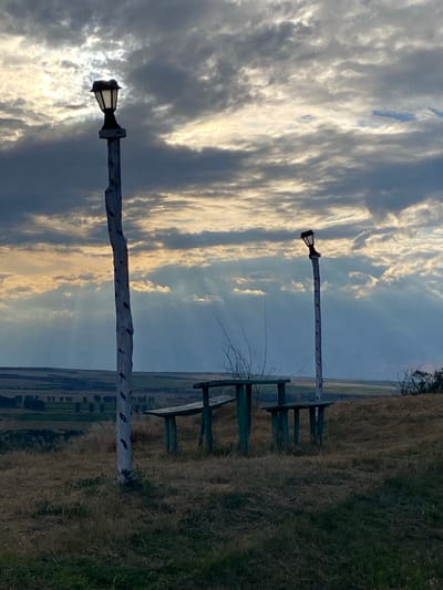 The image depicts an outdoor scene on a hill overlooking a rural landscape in Horești, Moldova. In the foreground, a rustic wooden picnic table with benches is positioned on dry grass and vegetation. Two tall, weathered wooden posts, each topped with a lantern, stand near the table. The sky above is dramatic with a mix of dark, heavy clouds and rays of sunlight breaking through, suggesting either dawn or dusk. The background shows rolling hills with fields and scattered trees. There are no people or animals visible in the scene.