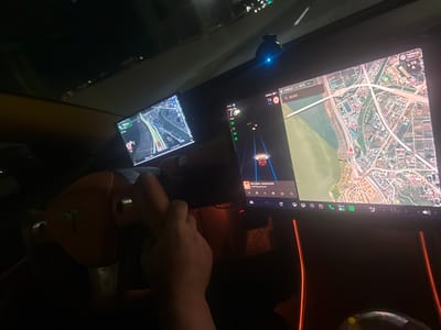 Here is a forensic description of the image:
**Scene:** The image is taken from inside a vehicle, likely a Tesla, at night.
**People:** A person's hands are visible on the steering wheel. The person's arms and torso are visible, but their face is not.
**Subjects:** Two displays are visible on the dashboard: one on the left showing a navigation map of the road and surrounding landscape and one main display on the right showing the car's driving and safety metrics. The main display is also showing a navigation map, the song "Harry Styles-American Girls," and car information (speed 73-74km/h), and environmental data (19°C).
**Objects:** The steering wheel features the Tesla logo. Other objects present are the dashboard, a rearview camera, and car interior details.
**Environment:** The scene is dark with interior lights illuminating the dashboard and screens. The screen displays indicate navigation in the area of Gimpo-si, South Korea, specifically showing "김포한강신도시 교차로" (Gimpo Hangang New City Intersection) and "신가지충전소" (Singaji charging station) on the main display. The secondary map shows the road ahead. The exterior environment is not clearly visible due to the darkness.