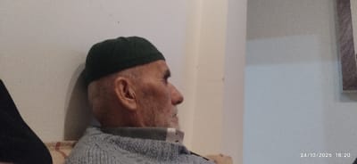 The image, captured on 24/10/2025 at 18:20, displays an elderly male seated indoors in Mu'adammiyat ash-Sham, Syria. The individual is presented in profile, facing right, wearing a dark green skullcap and a grey, possibly knitted, long-sleeved garment over a white-collared undershirt. He has a short, white beard. The immediate background is a light-colored, plain wall, with a partial wooden frame visible on the far right. A patterned fabric, indicative of upholstery or a cushion, is visible beneath the subject. A dark, indistinct object is partially visible at the far left of the frame.