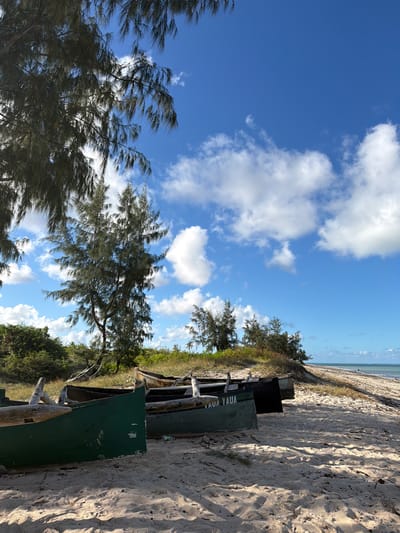 The image shows a beach scene, possibly in Pemba, Mozambique. The sky is blue with scattered white clouds. The beach is sandy and slopes down to the water in the distance. Several green boats are lined up on the sand, bearing text including "YAUA". Trees and other vegetation grow near the edge of the beach.