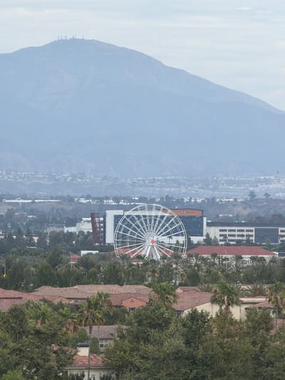 This image, captured in Irvine, California, presents a panoramic view of a cityscape nestled against a backdrop of hazy mountains. The foreground is dominated by a large, white Ferris wheel, its intricate spokes forming a striking geometric pattern against the muted sky. Several buildings with red-tiled roofs are visible, interspersed with lush green trees, including palm trees, suggesting a warm climate.
In the distance, a sprawling urban landscape stretches out, fading into a soft, atmospheric haze. The mountains in the far background appear muted and soft, their details obscured by the atmospheric conditions. The overall impression is one of a serene, perhaps overcast day. The presence of the Ferris wheel suggests a location that might be an entertainment district or a part of a larger complex. The architectural style of the buildings, with their red roofs and white facades, is typical of Southern California. No people are visible in the frame.
The image is oriented vertically, which may imply it was taken with a phone camera or has been rotated. The lighting suggests it is daytime, but the heavy haze or fog softens the light and obscures finer details, creating a diffused and tranquil atmosphere. The scene conveys a sense of scale, contrasting the man-made structures with the natural landscape.