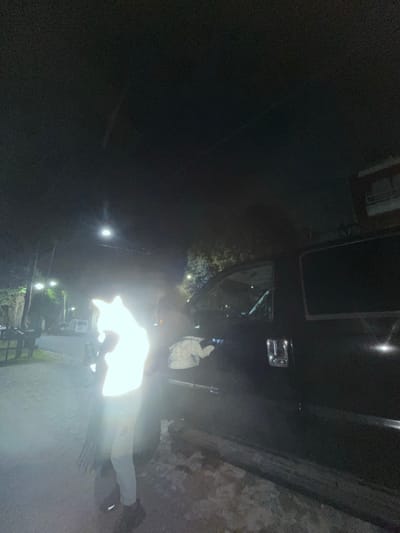 The photograph appears to be taken at night in what may be Olivos, Argentina, though this is not conclusively confirmed in the image. A person stands on a paved surface, possibly a sidewalk or road, illuminated by a bright light source that obscures their features. The upper part of a dark-colored vehicle, possibly a car or truck, is visible above and to the side of the person. In the background, streetlights are spaced along a street or walkway lined with greenery, likely trees or shrubs. A brick wall is visible on the right side of the image. The photograph is somewhat blurry, and significant portions are shrouded in darkness.