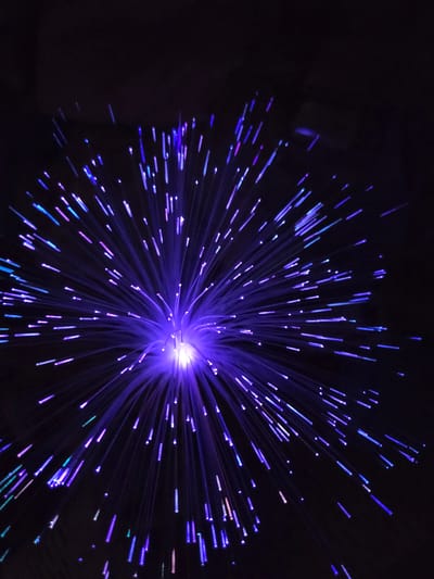 A close-up image shows a fiber optic lamp against a dark background. The light source is concentrated in the center, emitting numerous strands of light that radiate outwards. The strands are primarily blue and purple, with some hints of pink and green. The image lacks identifiable features linking it to Cherkovitsa, Bulgaria, or any specific location. There are no people or actions visible in the image.