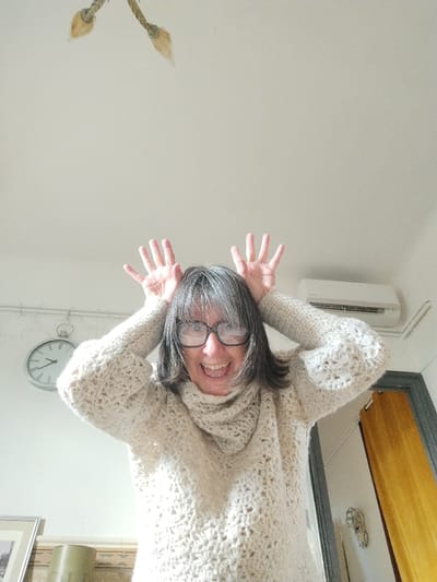 The scene depicts an interior room in Montélimar, France. An adult female, with grey and dark hair and dark-framed glasses, is centered in the image from approximately the waist up. She wears a light-colored, possibly off-white, chunky knit cowl-neck sweater. Her expression is a broad smile with an open mouth, revealing teeth. Both hands are raised to the sides of her head, with fingers splayed upward. The background consists of plain, light-colored walls and ceiling. Visible objects include a silver-toned, circular wall clock on the upper left wall, indicating a time near 1:55. A white, wall-mounted split-system air conditioning unit is positioned on the upper right wall. A decorative ceiling light fixture with two light-toned, leaf-shaped elements is partially visible at the top center. The bottom left shows a portion of a framed image and a cylindrical object on a surface with a decorative patterned border. A dark-framed doorway or opening is partially visible on the far right.