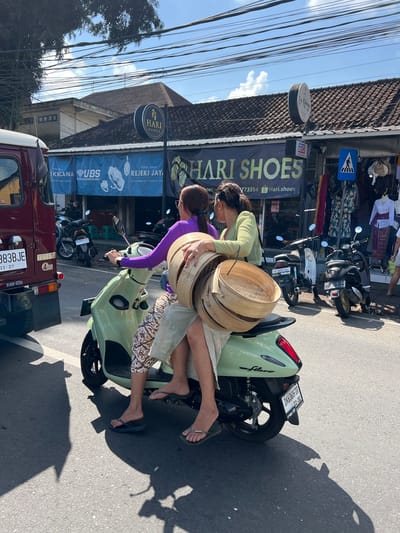 Two women are riding on a light green scooter on a paved street. The woman in the front is driving and is wearing a purple long-sleeved shirt and patterned pants. The woman in the back is wearing a green long-sleeved shirt and a light-colored skirt. She is holding a stack of round, woven bamboo containers. The scooter is carrying them past a row of shops. One shop has a banner that reads "HARI SHOES". There are other scooters and a red truck parked on the street. Overhead, there are numerous electrical wires. The scene appears to be in Abiansemal, Indonesia, given the presence of the "HARI SHOES" store which is a known establishment in that location. The sky is blue with some clouds, and the sun is casting shadows on the street.