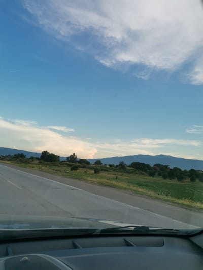 The image captures a view from inside a vehicle looking out at a rural landscape. A paved road with white lane markings curves to the left in the foreground. To the right of the road, a grassy embankment slopes upward, dotted with trees and some low-lying vegetation. In the middle ground, a range of rolling hills covered in trees forms a natural barrier. Beyond the hills, a vast expanse of blue sky stretches across the frame, punctuated by scattered white clouds, some of which appear wispy and elongated, possibly indicating the presence of high-altitude clouds or contrails from an aircraft. The overall scene suggests a journey through a picturesque rural area, possibly on the outskirts of Krivina, Bulgaria, given the context. The weather appears to be fair with clear skies.