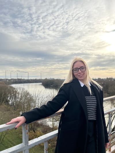 A blonde female, wearing dark-rimmed glasses, a white collared shirt, a dark horizontally striped sweater, and a black belted overcoat, stands in the foreground, facing slightly towards the viewer with a faint smile. Her left hand rests on a visibly rusted, light-colored metal railing. The scene provides an elevated perspective over the Moskva River in Voskresensk, Russia, which flows through the mid-ground. The riverbanks and small islands within the river are lined with bare deciduous trees, indicative of a colder season. In the distant background, at least three tall industrial chimneys are prominent, actively emitting plumes of smoke or steam. Scattered low-rise buildings are visible adjacent to the industrial structures. The sky is heavily overcast with multiple layers of clouds, exhibiting a diffused bright area in the upper right quadrant, suggesting a late afternoon or early evening light.