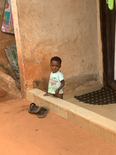 The scene is set in Otuocha, Nigeria. A young child, appearing to be a toddler, stands looking directly forward from within an entrance or recessed area. The child has dark skin and short dark hair, and wears a light green short-sleeved t-shirt featuring "WORLD CUP 2022" text and a graphic. The child's left hand rests on a low, light-colored tiled ledge that forms the threshold of the entrance. In the foreground, on a reddish-brown dirt ground, two dark-colored open-toe sandals are positioned to the left of the ledge. The background consists of a textured, light brown wall exhibiting signs of wear and discoloration. To the far left, a colorful patterned mat hangs partially visible on the wall, with another folded item below it. To the right, a dark wooden doorframe is visible, with a dark, patterned doormat placed on the floor inside the threshold. The area is illuminated by natural light.