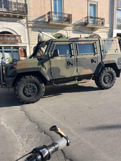 The image depicts a camouflaged military vehicle parked on a city street. The vehicle is positioned in front of a building with wrought iron balconies and a cafe called "Caffè Alighieri." A bicycle handlebar is visible in the foreground. The location is Galatina, Italy, as identified by the cafe name. A person in a blue shirt is standing near the front of the vehicle. The surrounding environment appears to be an urban setting with typical European architecture.